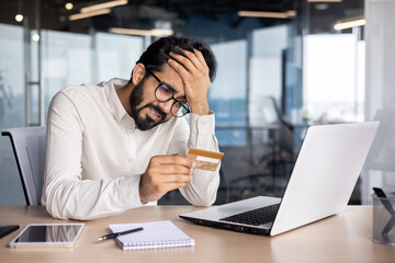 Frustrated young muslim man sitting in office at desk in front of laptop and looking at credit card, holding head upset