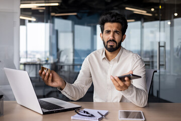 Portrait of a worried and upset young Indian man sitting at a desk in the office, holding a phone and a credit card in his hands and waving his hands to the camera in frustration - obrazy, fototapety, plakaty