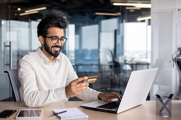 Smiling muslim male businessman sitting at desk and working in office on laptop and using credit card - obrazy, fototapety, plakaty