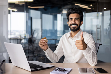 Portrait of a smiling young Indian man sitting in the office at the desk, holding a credit card in his hand and showing the super sign with his finger to the camera