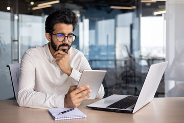 Concentrated and serious young Indian man working in office at desk with tablet - obrazy, fototapety, plakaty