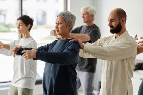 Side view of bearded male trainer correcting shoulder position of gray-haired senior woman during qigong exercise
