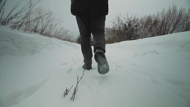 Legs of a man walking on snow with footprints on a snowy day. Close-up of the legs of an unrecognizable man in black leather boots walking on a snowy road in winter.
