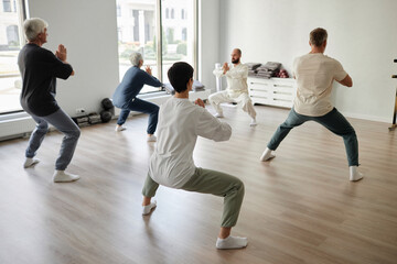 Back view at senior qigong students doing concentration exercise in half-squat position, male trainer showing prayer pose - obrazy, fototapety, plakaty