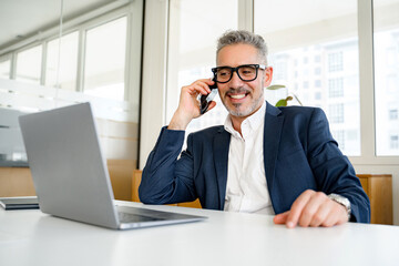 Confident mature 50s businessman talking on phone sitting at the desk with laptop in office,...