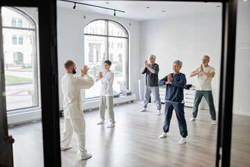 Wide angle shot of mixed group of senior qigong students repeating prayer pose exercise after male...