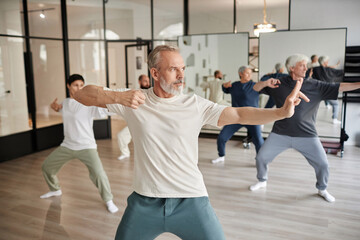 Fototapeta na wymiar Side view of elderly people in gym, focus on bearded senior man posing as archer shooting during qigong lesson in gym