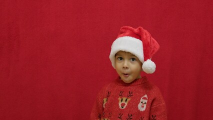 Funny face expression of a cute preschool boy with christmas sweater and santa claus hat, isolated on red with copy space. High quality photo