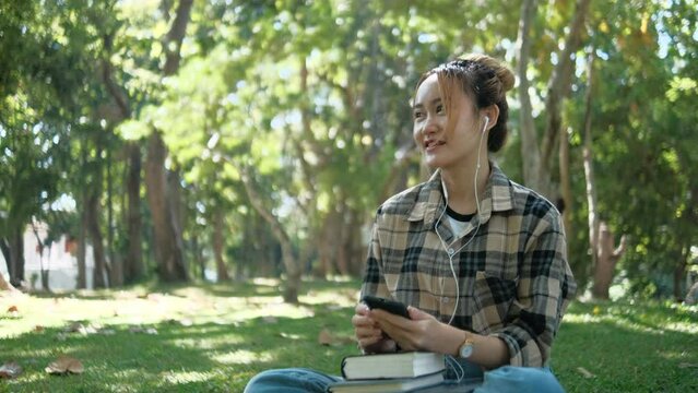 A young woman sitting in a public park uses her phone and listens to audio on her headphones.
