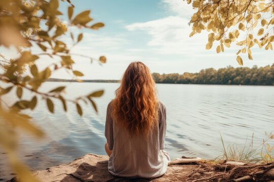 Back view of a redhead woman sitting near lake on the shore. Digital detox concept