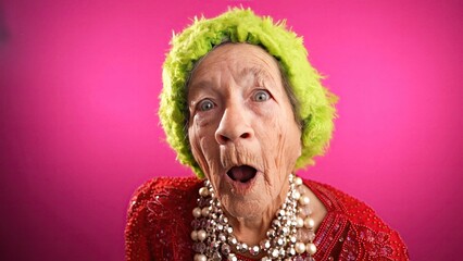 Laughing funny elderly woman saying WOW having great success with green hat isolated on pink...