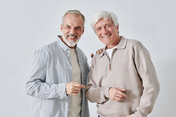 Waist up shot of blue-eyed bearded senior man dressed in striped shirt placing hand on shoulder of white-haired senior male friend, both laughing and looking at camera