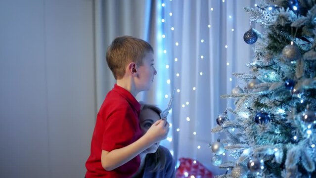 Mother and boy are enthusiastically decorating the Christmas tree, hanging balls and toys. The lights shine magically, and the festive mood grows, turning every twig into a fairy-tale image. 