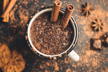 Homemade spicy hot chocolate drink with cinnamon stick, star anise, grated chocolate in enamel mug on dark background with cookies, cacao powder and chocolate pieces, top view