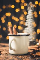 Homemade spicy hot chocolate drink with cinnamon stick, star anise, grated chocolate in enamel mug on dark background with cookies, cacao powder and chocolate pieces, Christmas lights bokeh