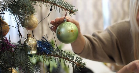 Mom reaches out her hand in the branch of the Christmas tree and hangs a New Year's toy on it. Mom and children decorate the Christmas tree for the New Year. - obrazy, fototapety, plakaty