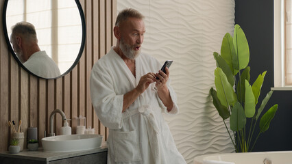 Old Caucasian man senior mature retired male in bathrobe in bath bathroom at morning looking mobile...