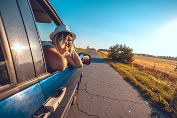 A beautiful woman driving a car enjoys the feeling of the wind in her hair and freedom. A woman looks out the open car window and the wind blows her hair. The concept of traveling by car. - obrazy, fototapety, plakaty