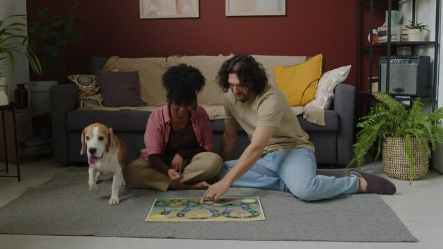 Young interracial couple with lovely beagle dog sitting together on floor in modern living room chatting and playing boardgame