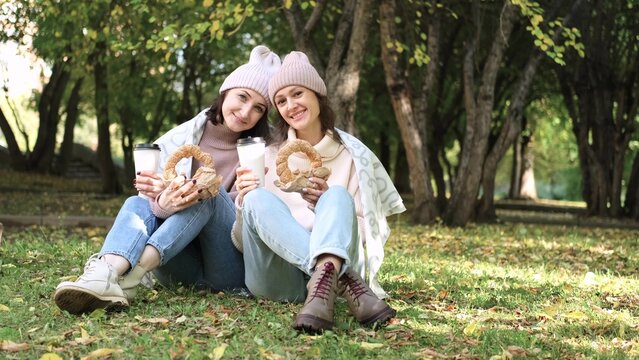 Two cute women in warm clothes sit hugging in an autumn park. Girlfriends enjoying beautiful weather, drinking tea or coffee, eating fresh bagels from the bakery