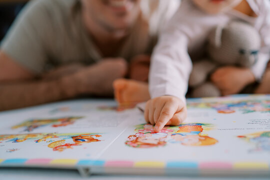 Little girl points her finger at a colorful picture in a book while sitting with her dad. Cropped. Faceless