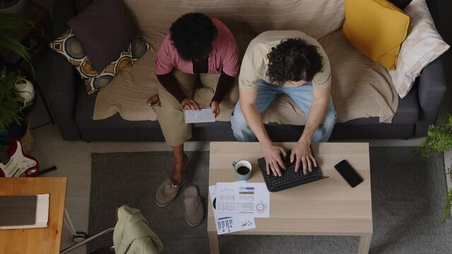Top view shot of young ethnically diverse married couple managing finances and counting bills, sitting together on sofa in living room using digital tablet and laptop computer