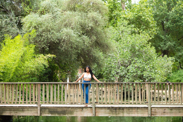 Fototapeta na wymiar Young and beautiful brunette woman and latin, leaning on the railing of the wooden bridge over the duck and fish pond in the park. The woman is smiling and happy and enjoying the day.
