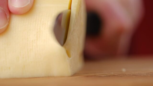 Close up of cheese being sliced at home in preparation for sandwich for lunchtime. Unknown young woman cuts piece of cheese with knife on wooden board