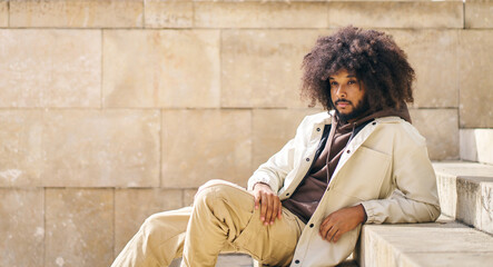 Young man with afro hair sitting on the steps of a quiet square in the city on a sunny day