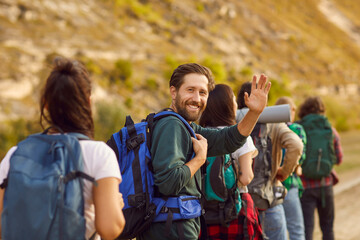 Portrait of a happy smiling bearded man waving hand looking cheerful at camera while trekking with backpack during a walking tour of the mountains with a group of his young friends. - obrazy, fototapety, plakaty