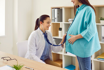 Pregnancy checkup. Female obstetrician examines pregnant woman's abdomen with stethoscope in...