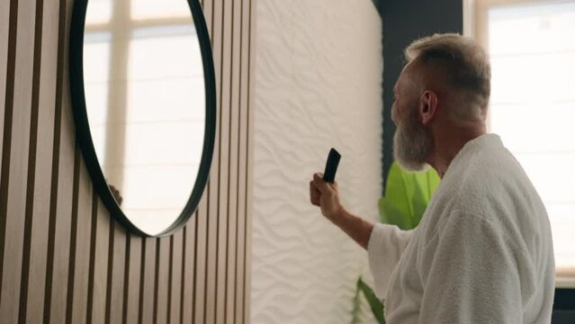 Retired old Caucasian man senior mature male grandpa in bathroom looking at mirror reflection having fun enjoy morning beauty routine singing song in hair brush comb like microphone karaoke sing funny