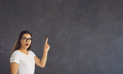 Shocked surprised woman presents incredible proposal by pointing her finger at copy space. Young brunette woman with glasses standing on gray background looks at camera with wide open eyes and mouth.