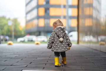 A cute stylish two-year-old toddler girl kid in a gray coat and yellow boots walks outdoors in the...