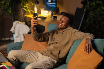 Delighted attractive young African Aerican guy blogger writer, crafting articles or essays on his laptop while lounging on the sofawearing casual outfit.