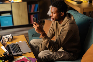 Concentrated serious African dark skinned black video production assistant, using his phone to communicate with colleagues and capture behind-the-scenes footage while editing raw footage on laptop. - obrazy, fototapety, plakaty