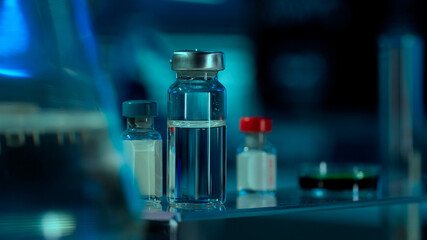 Close up of glass vials with clear liquid and petri dish on table in research lab or hospital. Vials with clear liquid on blurred background. Concept of laboratory research, health care.
