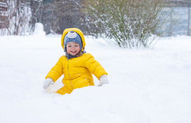 Fototapeta na wymiar happy little child in yellow winter clothes playing in snow under falling snowflakes outdoors