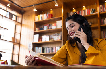A beautiful young girl business woman or student talking on phone while working or studying in school or collage library or in corporate office sitting with books