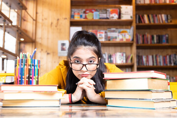 A beautiful young girl business woman or student wearing glasses spectacles of eyeglass seating with pile of books working studying in school collage library or in corporate office sitting with books