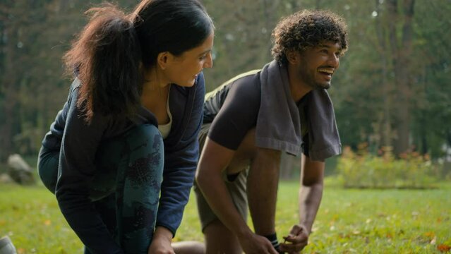 Two sporty people fixing sneakers tie shoelace of shoes together talking conversation couple Arabian man sportsman and Indian woman sportswoman prepare to run fitness sport workout walking in park