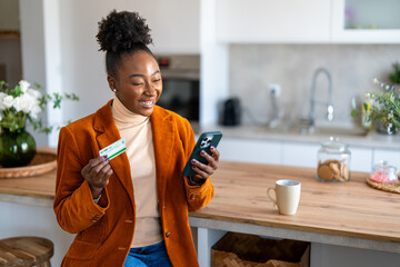 Charming young black woman looking at smart phone screen holding credit card, using mobile phone...