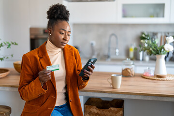 Serious young black woman wearing business casual clothes sitting at home while using mobile phone and credit card.