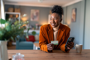 Beautiful African businesswoman sitting at home office using smart phone and credit card. - obrazy, fototapety, plakaty