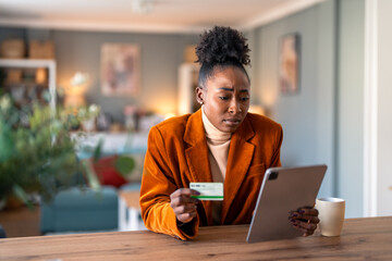 Worried young adult woman holding credit card while checking electronic bank account over digital...