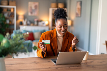 Happy young female shopper holding credit card, using laptop doing online banking transaction while sitting at desk in home office during the day.