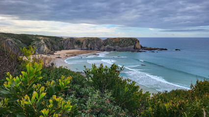 Zambujeira do Mar over the sea shore with ocean waves, cliffs and sand dunes covered by green vegetation red leaves of sour fig, sunny day, clear blue sky. Rota Vicentina coast, Portugal.