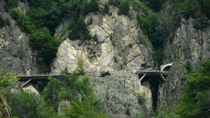 A road passing stony mountainsides by a tunnel and by a suspended bridge. The vertical cliffs are...