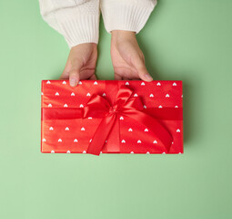 Female hands holding a gift box with a red ribbon on a green background. View from above