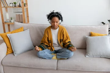 Poster Yoga mindfulness meditation. Young healthy african girl practicing yoga at home. Woman sitting in lotus pose on couch meditating smiling relaxing indoor. Girl doing breathing practice. Yoga at home © Юлия Завалишина